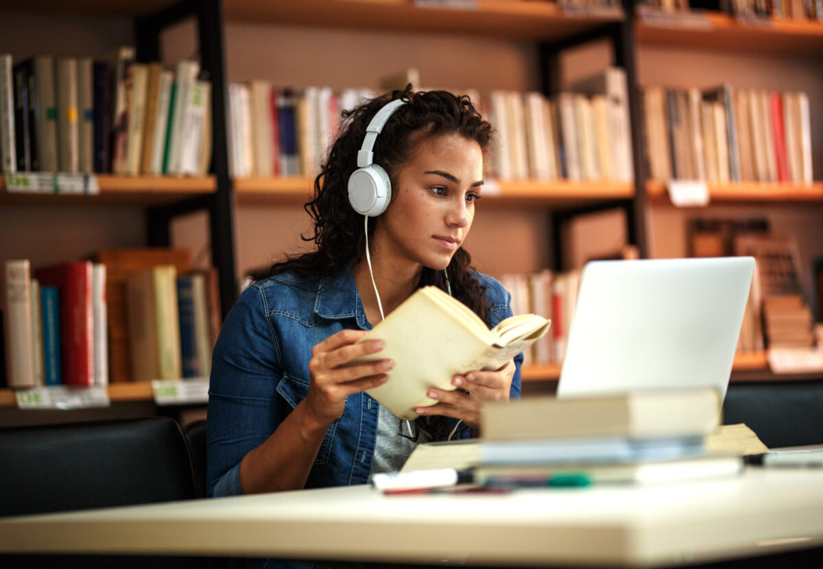 Female student in library, listening to music, studying on her computer, book in hand