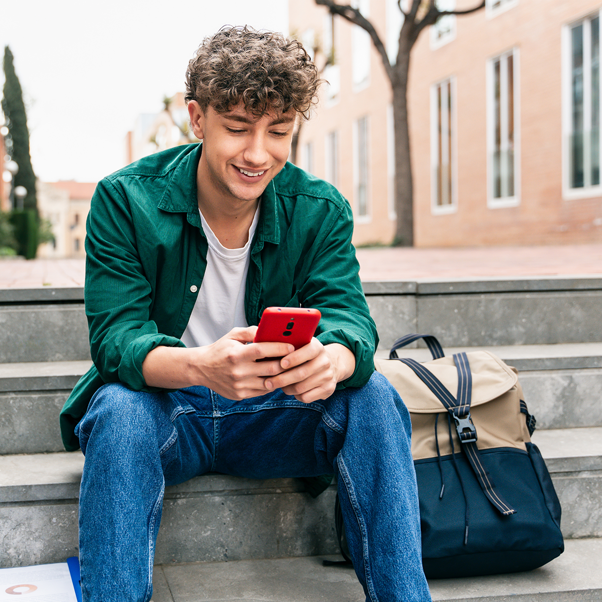 Male student sits on steps outside campus and looks down at his phone.