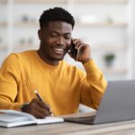 Happy african american young man freelancer or businessman working from home, sitting at table in front of modern laptop, having phone conversation, taking notes and smiling