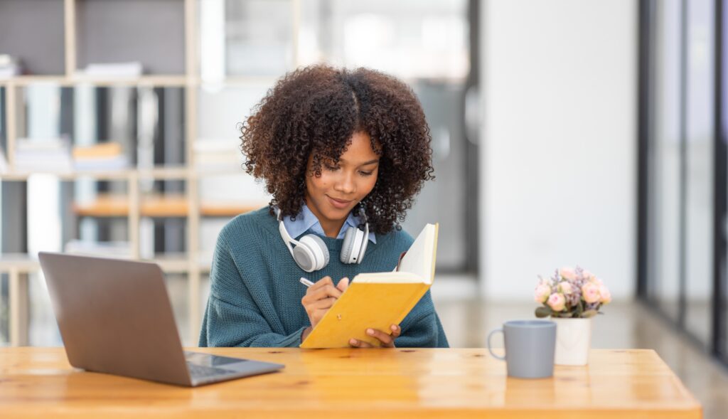 Cute african american female student wearing headphones with afro dreadlocks, studying remotely from home, using a laptop, taking notes on notepad during online lesson, e-learning concept,