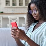 Young happy smart African black student girl businesswoman in glasses sitting on sofa holding using cell mobile phone apps reading e book, surfing social media, online chatting in apartment.