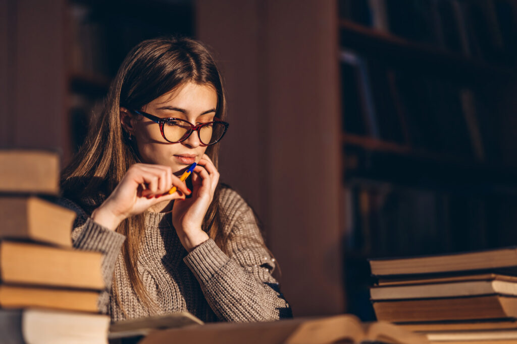 Female student sits at a table in the library with a pile of books.